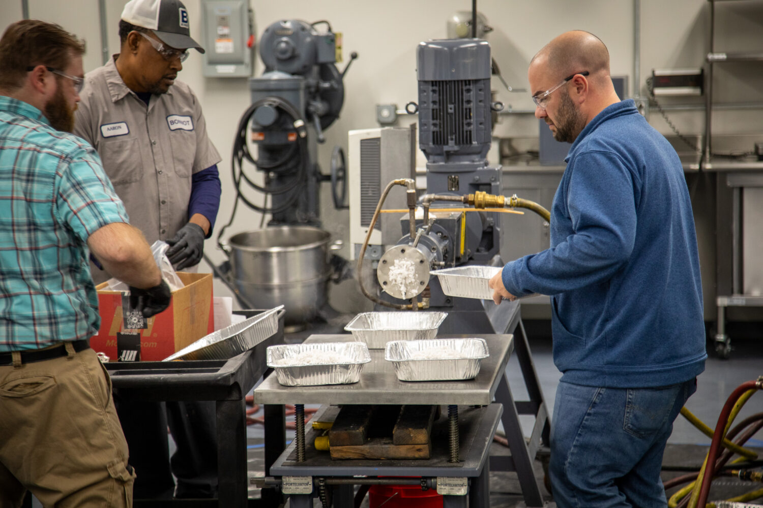Three men work in an industrial kitchen, preparing aluminum trays on a table. Machinery—including equipment from a single screw extruder manufacturer—and various tools are visible in the background. All men wear safety glasses; two wear work uniforms and gloves.