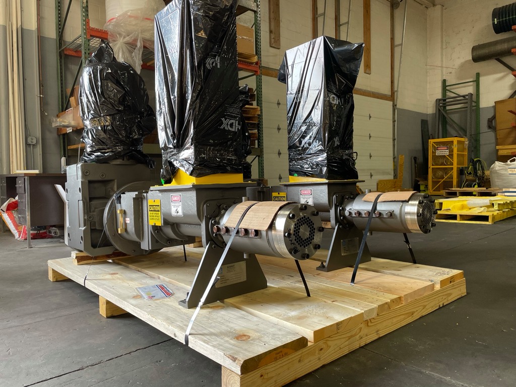 Three large industrial machines are mounted on a wooden pallet in a warehouse. The upper parts are covered with black plastic bags, and various warning labels are visible on the machines. Shelving and tools are in the background.